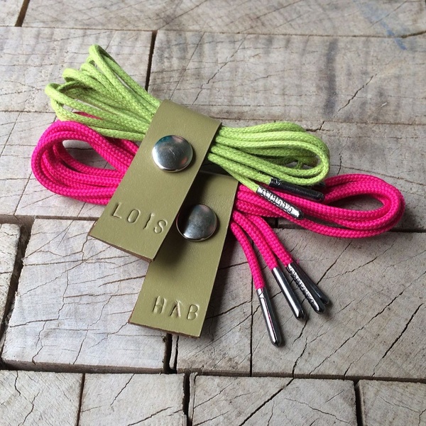 A close-up studio shot featuring two pairs of vibrant shoelaces with branded leather tabs on a textured wooden background.