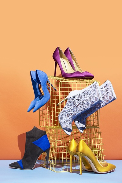 A sophisticated product hero display featuring multiple high heel styles arranged on a yellow wire grid against a vibrant orange backdrop.