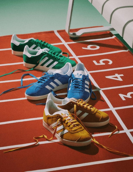 A high-angle 4:5 shot of three pairs of suede sneakers placed on a stylized red running track with lane numbers, emphasizing color and texture.