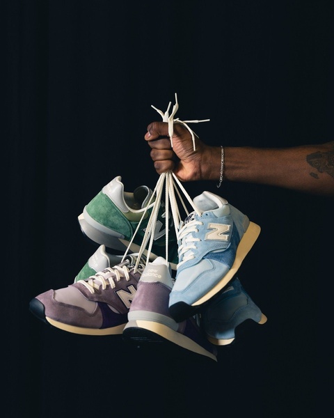 A high-contrast, minimalist studio shot of a hand holding multiple pairs of running shoes by their laces against a solid black background.