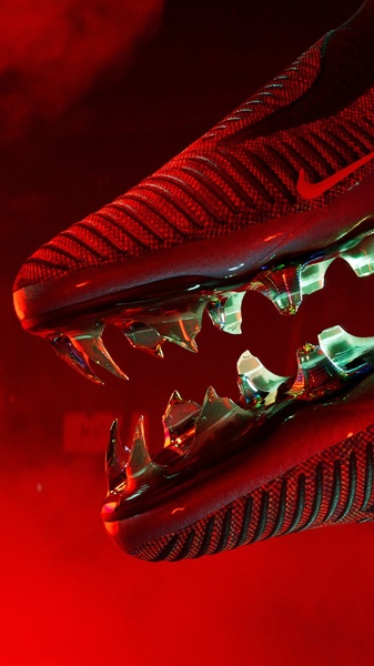 A dramatic, macro-style close-up shot of a high-performance soccer cleat against a deep red, smoky background, emphasizing the aggressive traction and sleek texture.