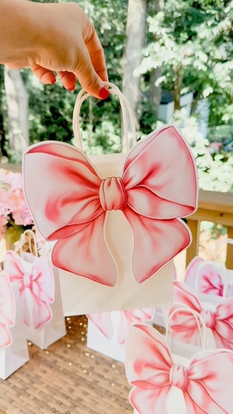 A close-up shot of a hand holding a clean white paper gift bag featuring a large, illustrated pink bow. The scene highlights simple, elegant product packaging.