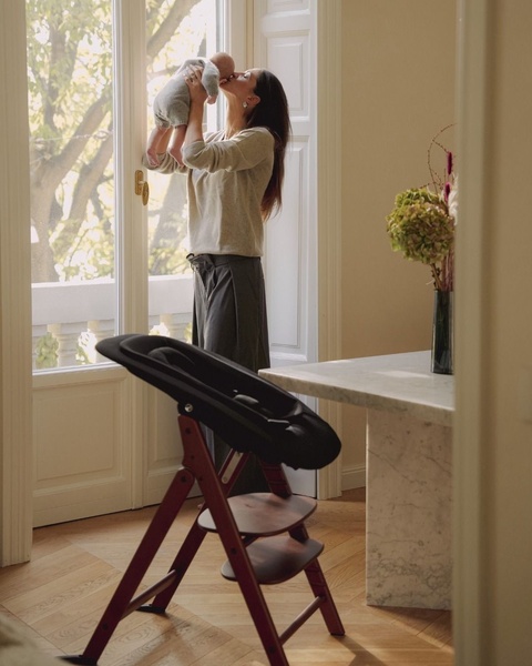 A high chair presented in a serene home setting, with a mother interacting with her infant in the background to convey emotional warmth and high-end lifestyle.