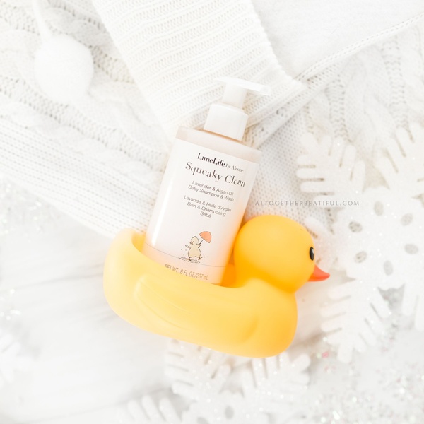 A high-key, bright, and clean product shot of a baby shampoo bottle nestled with a yellow rubber duck, set against a soft, textured fabric background.