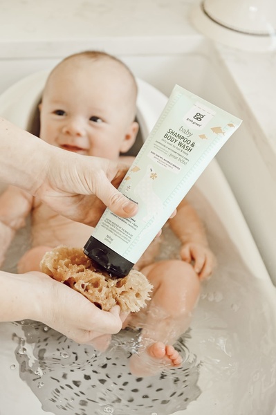 A close-up lifestyle photograph showing a baby in a tub while a hand applies gentle shampoo and body wash using a natural sponge.