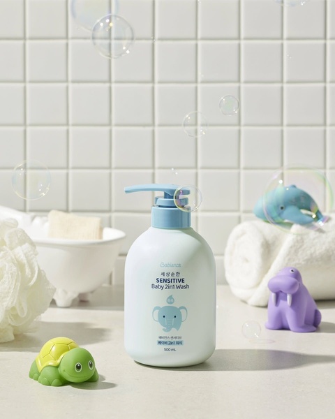 A clean, bright studio shot of a Baby 2in1 Wash bottle surrounded by playful bath toys and soap bubbles against a tiled background.