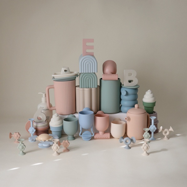 A high-end, clean product hero shot featuring a collection of pastel silicone baby dishware items arranged in a tower-like, symmetrical structure.