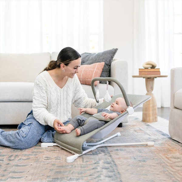 A natural, high-key lifestyle photograph featuring a mother and baby interacting with a neutral-toned baby bouncer in a home setting.