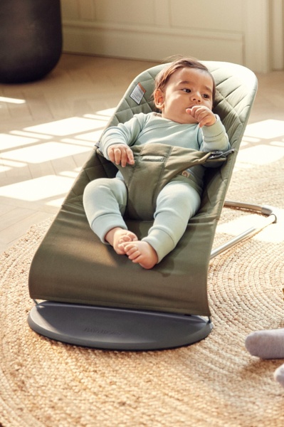 A high-quality lifestyle shot featuring a baby in a neutral-toned bouncer on a natural rug, emphasizing comfort and premium design.