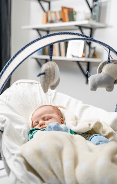 A soft-focus lifestyle image of an infant sleeping peacefully in a modern, neutral-toned baby bouncer with hanging mobile toys.