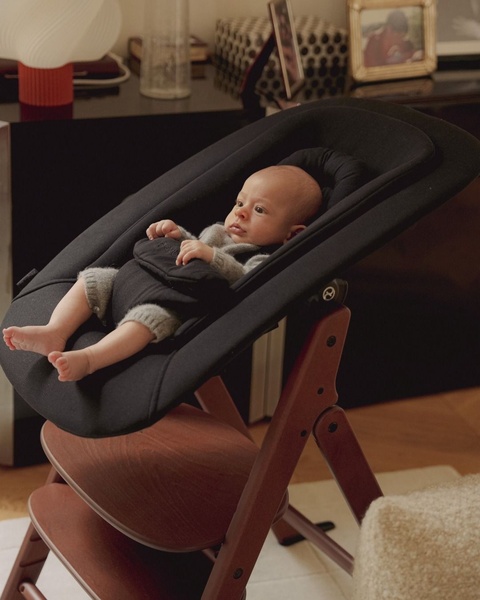 A high-quality, 4:5 lifestyle image featuring a baby resting in a dark-toned bouncer within a warm, domestic living space.