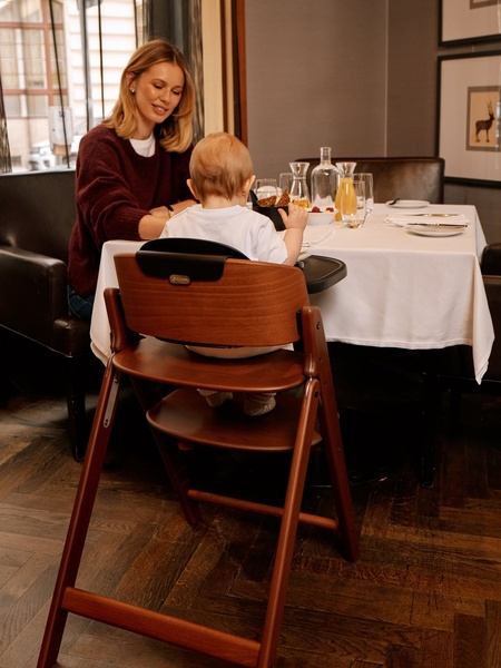 A high-quality lifestyle shot featuring a wooden high chair at a dinner table, focusing on the product's premium aesthetic and integration into home life.