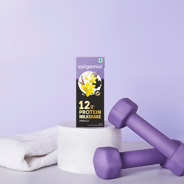 A minimalist studio shot of a protein milkshake carton placed on a pedestal with dumbbells and a towel, set against a soft purple backdrop.