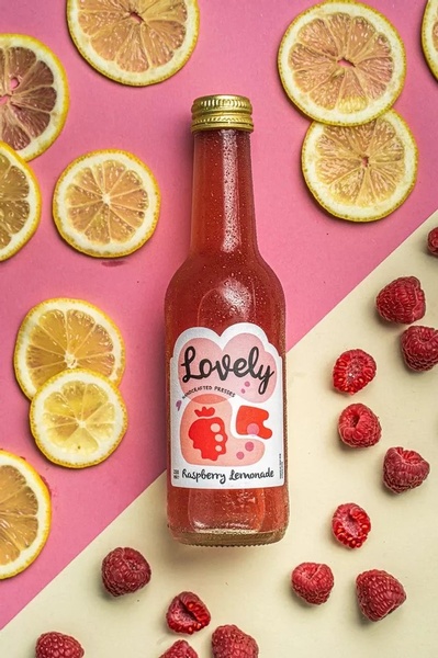 A high-contrast, symmetrical flat-lay style product photograph of a bottle of raspberry lemonade surrounded by sliced lemons and scattered raspberries.