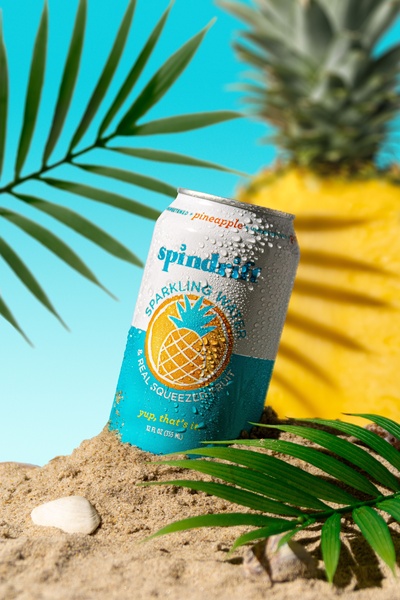 A high-contrast product hero shot featuring a can of sparkling water in sand surrounded by fresh pineapple and palm leaves.