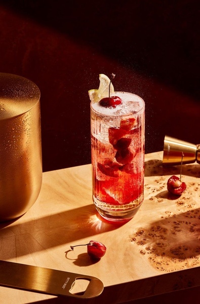 A high-contrast, professional-grade photograph of a red cocktail in a textured glass with a cherry and lemon garnish, set against a deep red background.