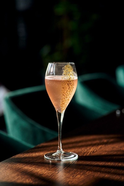 A high-end, studio-lit photo of a pink cocktail in an elegant flute glass, featuring a signature gold-glitter garnish and dark, moody background.