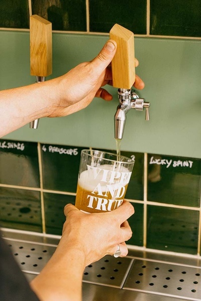 A close-up shot of a hand pouring fresh craft beer into a branded glass from a wooden tap handle.