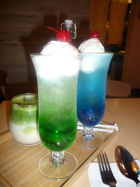 This creative showcases two vibrant soda floats topped with whipped cream and cherries in a casual cafe setting.