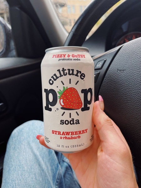 A close-up, first-person perspective shot of a Strawberry and Rhubarb probiotic soda can held in a hand while inside a vehicle.