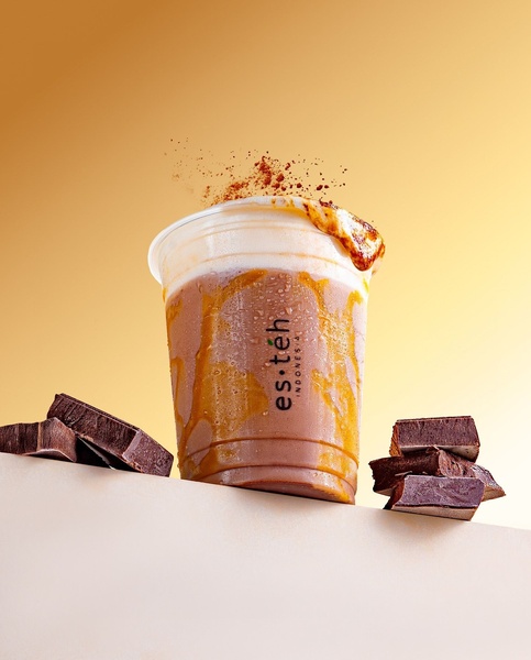 A high-contrast studio shot of an iced chocolate drink in a transparent cup topped with whipped cream and chocolate powder, flanked by dark chocolate bars against a warm gradient backdrop.