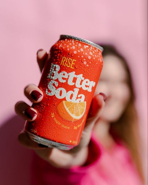 A close-up, high-angle shot featuring a condensation-covered orange soda can held toward the camera against a soft pink backdrop.