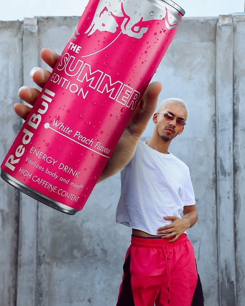A high-impact perspective shot where a hand holds a can of energy drink directly to the camera lens, dwarfing the model in the background to emphasize the product.