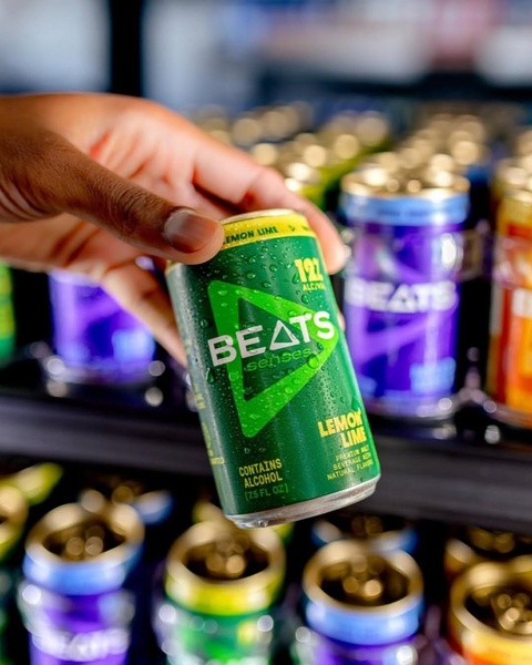 A close-up shot of a hand holding a condensation-covered green malt beverage can in a refrigerated display setting.