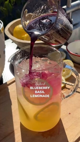 A top-down, sensory-focused shot of colorful, fruit-infused liquid being poured into a pitcher filled with ice and lemon slices.