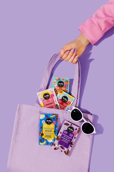A top-down, high-fashion style shot featuring multiple seed butter pouches held in a matching tote bag against a solid purple backdrop.