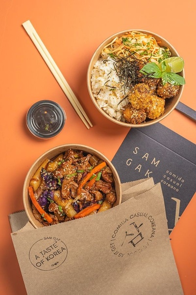 A high-quality, professional top-down photograph of Korean rice bowls, takeout packaging, and chopsticks on a warm-toned background.
