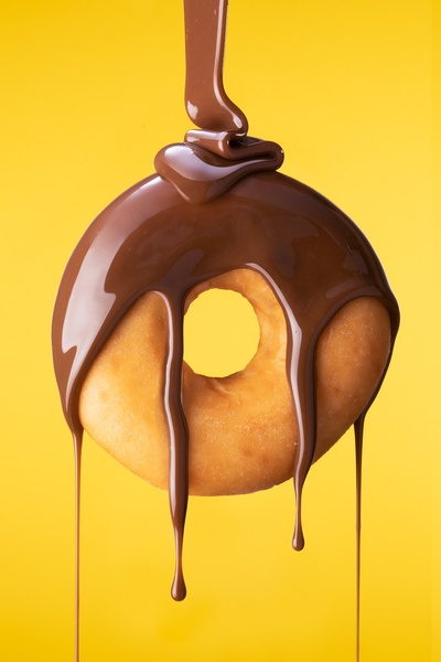 A close-up, high-definition shot of a plain donut being coated with a thick, cascading layer of liquid chocolate against a vibrant yellow background.