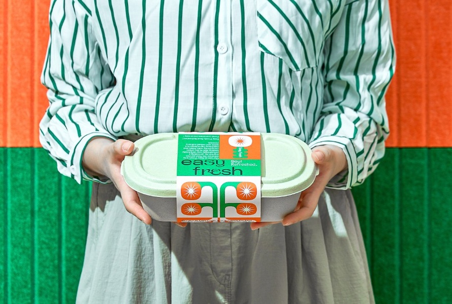 A close-up, symmetrical studio shot featuring a person holding a sustainable food container with bold orange and green branding.