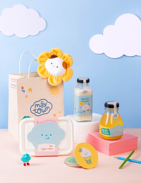 A bright, playful product hero shot featuring bottled beverages, a branded tote bag, and custom stationery against a light blue backdrop.