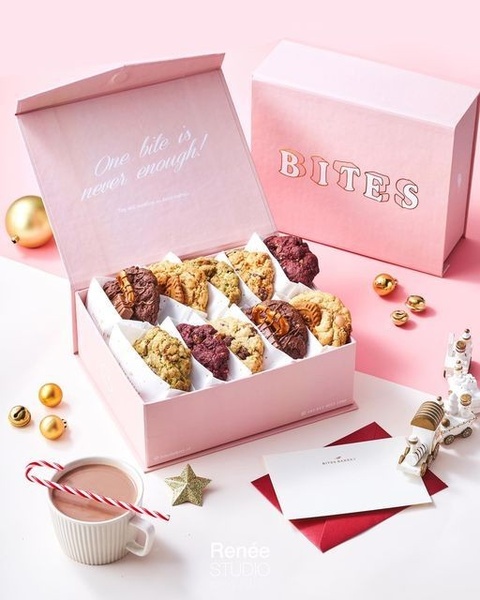 A top-down studio shot of an open luxury cookie gift box featuring assorted gourmet cookies, decorative ornaments, and a handwritten card on a soft pink background.