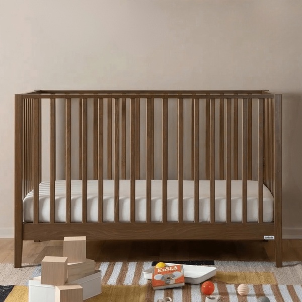 A high-quality, clean-shot product image of a wooden baby crib placed in a neutral-toned room with simple wooden building blocks in the foreground.