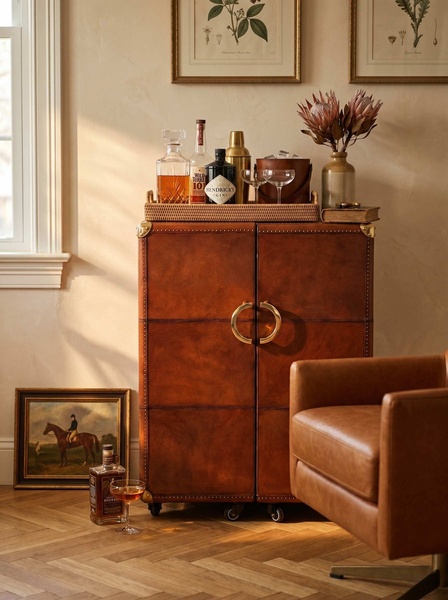 A sophisticated lifestyle shot featuring a premium leather bar cabinet, styled with glass decanters and botanicals in a warm-toned, elegant room setting.