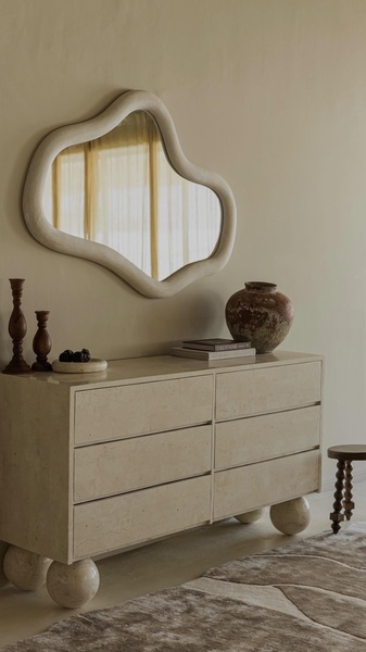 A high-end interior design shot featuring an organic-shaped wall mirror hanging above a stone-textured dresser in a minimalist, warm-toned room.