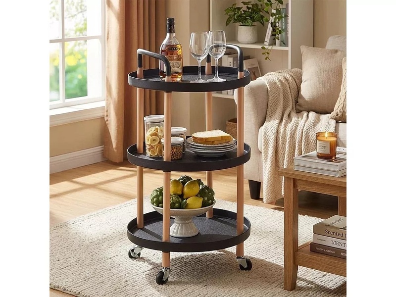 A high-quality lifestyle image of a three-tier black and wood round utility cart placed next to a sofa, styled with everyday home accessories.