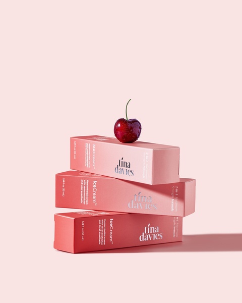 A clean, studio-lit shot featuring three tubes of cream stacked vertically with a single cherry resting on top against a soft pastel background.