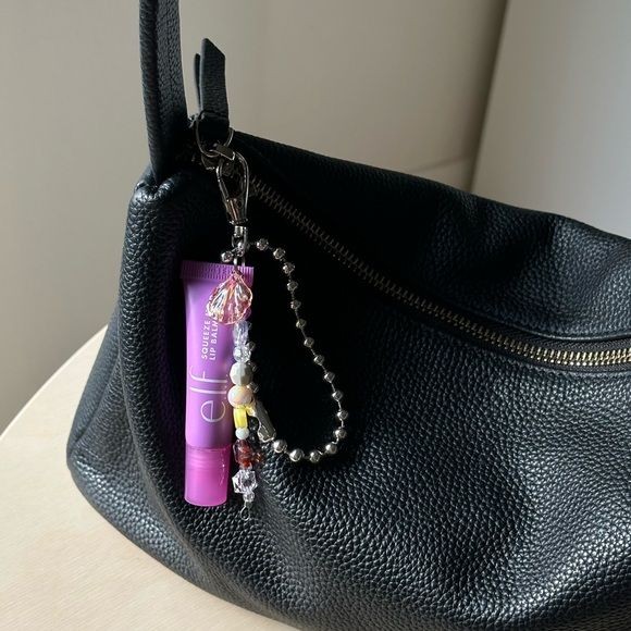 A close-up view of a purple lip balm tube attached as a keychain charm to a black textured handbag, emphasizing portability and lifestyle integration.