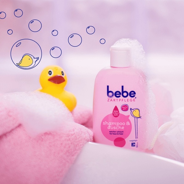 A close-up, high-key shot of a pink baby shampoo bottle sitting in a bathtub, surrounded by bubbles and a yellow rubber duck, creating a sense of gentle play.