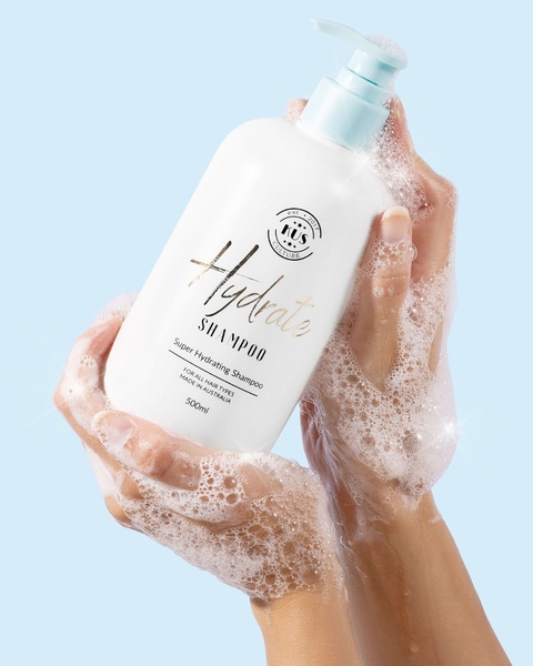 A close-up, high-key shot of a Hydrate Shampoo bottle held in sudsy, clean hands, emphasizing product texture and clarity.