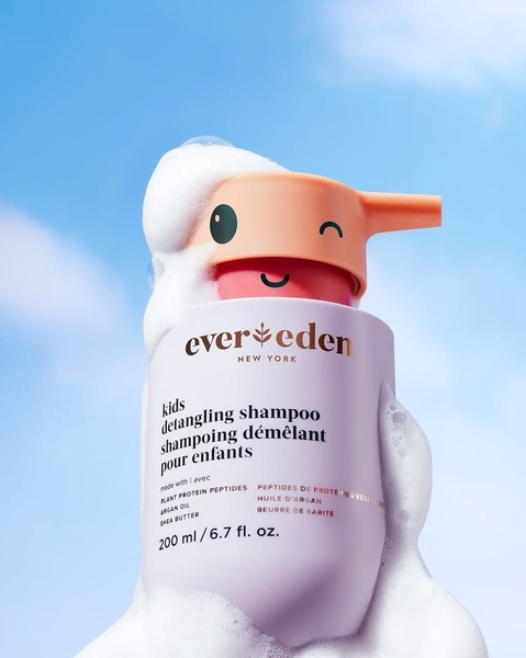 A close-up, high-key shot of a kids shampoo bottle emerging from a cloud of soap bubbles against a bright, clear blue sky background.