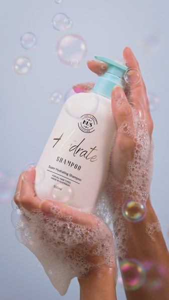A close-up, high-aesthetic shot of Hydrate shampoo being held in bubbly, hydrated hands against a soft blue background.