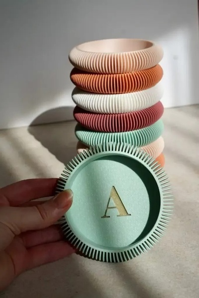 A close-up aesthetic shot of a personalized coaster held in front of a stack of ribbed, minimalist coasters in various pastel colors.
