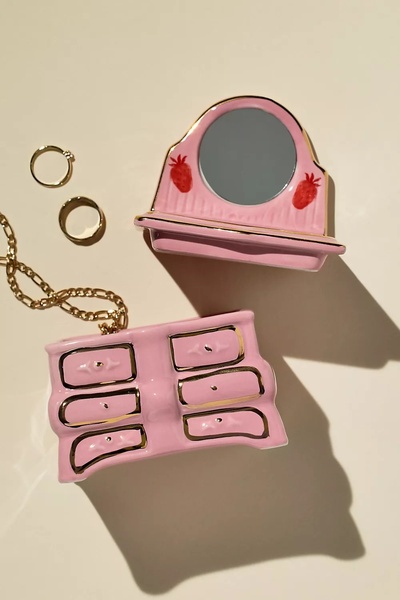 A high-angle product photograph of a pink, strawberry-themed miniature vanity mirror accompanied by golden rings on a neutral background.