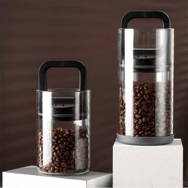 A high-contrast, clean product-hero shot showcasing two sizes of glass coffee bean containers with black handles on a white platform.