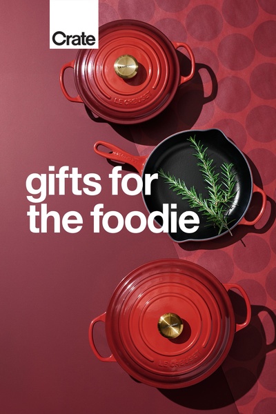 A top-down, clean product hero shot of two Le Creuset dutch ovens and a skillet, styled with fresh rosemary on a deep red background.