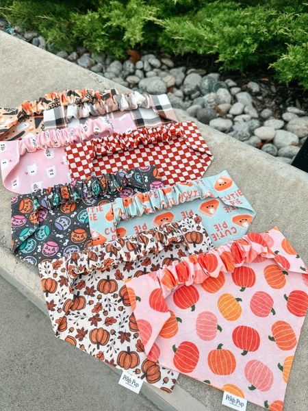 A high-angle flat lay showcasing a collection of autumn-themed dog bandanas featuring pumpkins, ghosts, and plaid patterns.